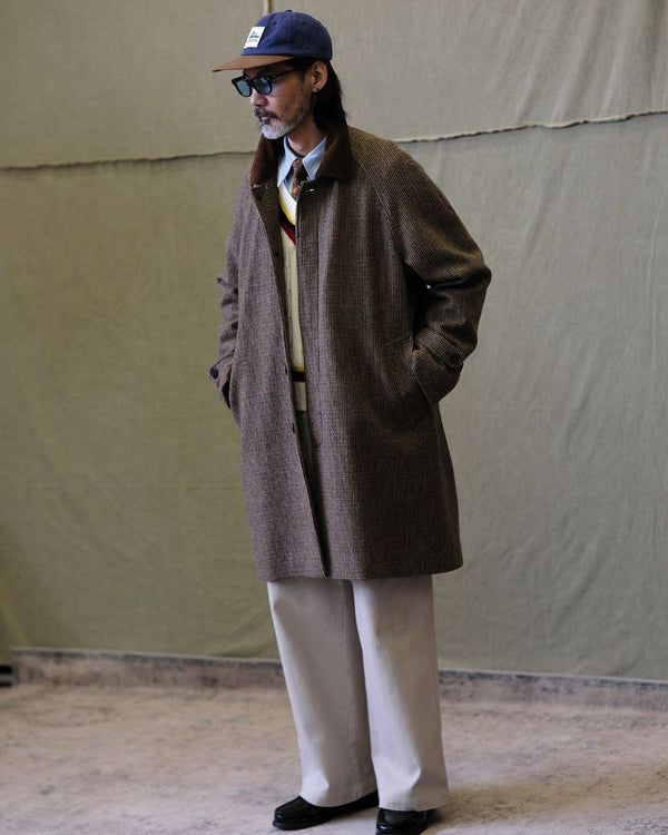 Man wearing a houndstooth balmacaan brown coat, beige pants, and a cap against a plain background