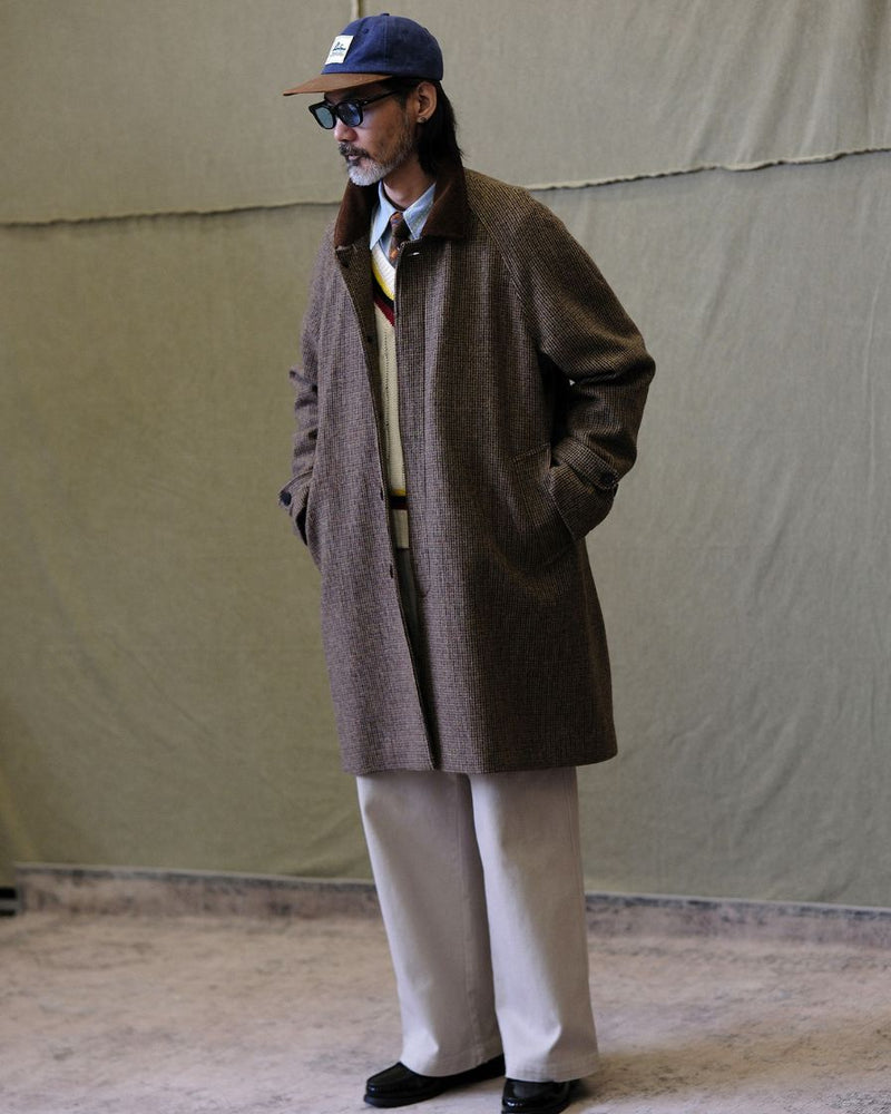 Man wearing a houndstooth balmacaan brown coat, beige pants, and a cap against a plain background