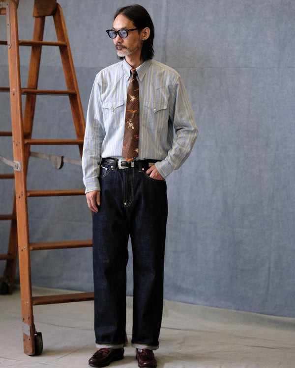 Man wearing a patterned shirt, tie, and dark pants standing in front of a gray wall with a wooden ladder.